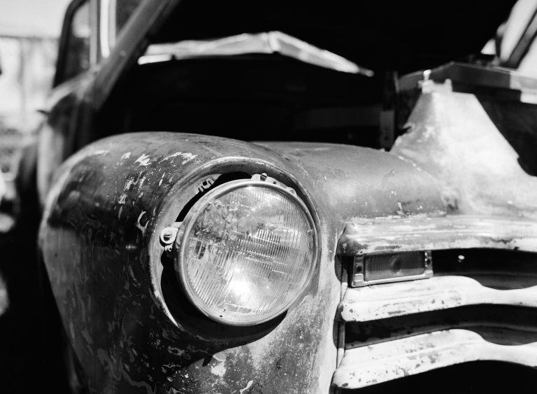 Close-up of the front of an old, weathered car with a round headlight and chipped paint. The hood is open, revealing part of the engine compartment in this striking black and white film photo. | Photo by Millworks Film Photography & Prints