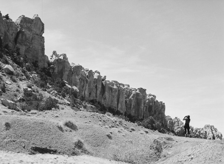 A person stands on rocky terrain gazing at a dramatic cliffside with jagged rock formations under a bright, clear sky in a striking black and white film photography landscape. | Photo by Millworks Film Photography & Prints