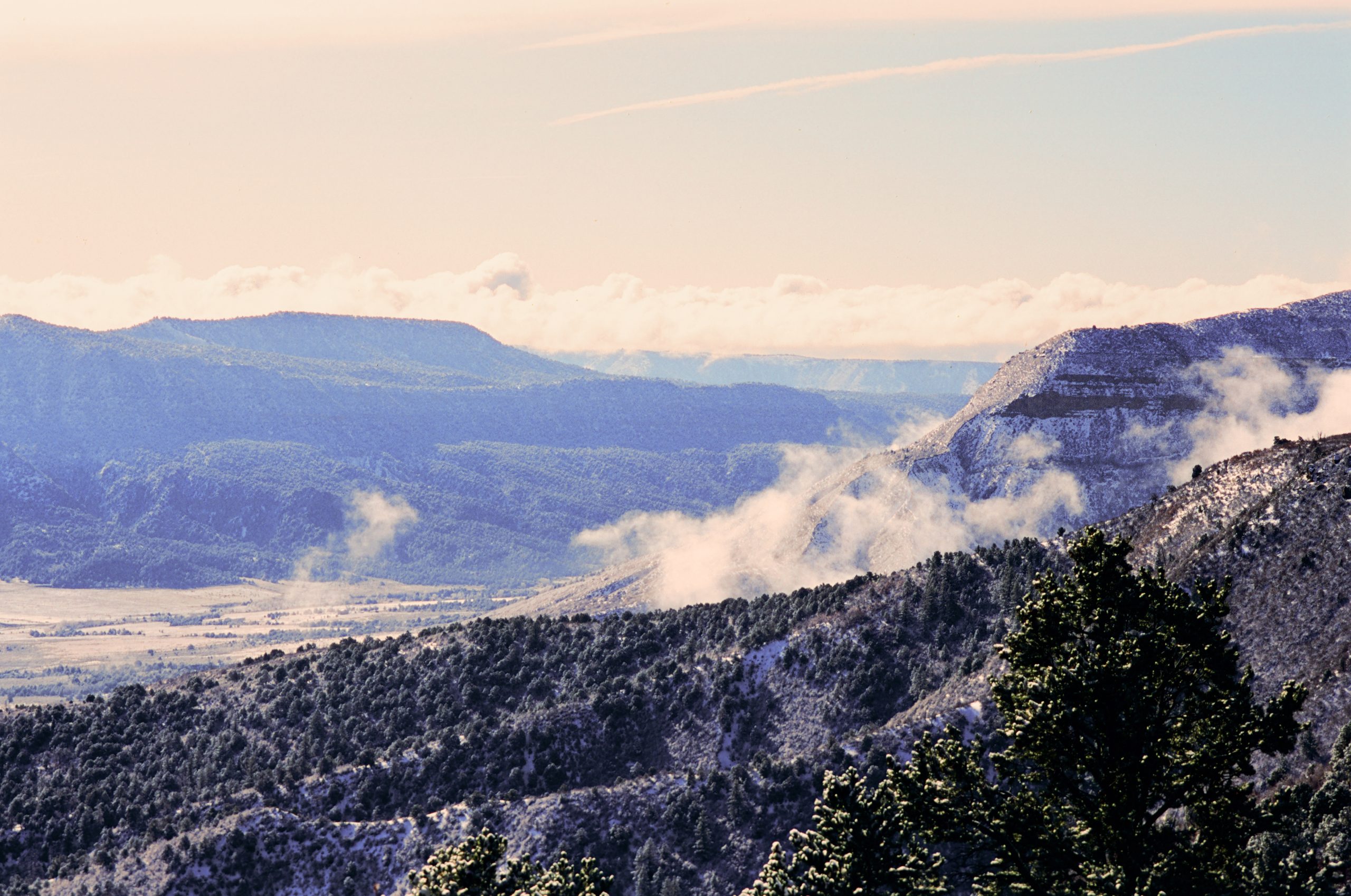 Snow-dusted mountains and hills with patches of evergreen trees, low clouds, and mist drifting through the valleys under a pale sky—captured in the timeless style of film photography. | Photo by Millworks Film Photography & Prints