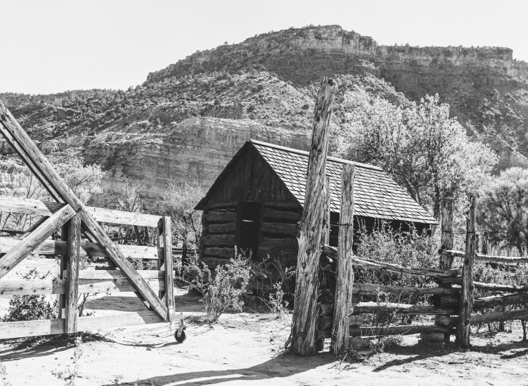 A black and white film photo of a rustic wooden cabin surrounded by a wooden fence, set in a dry, rugged landscape with rocky hills and sparse trees in the background. | Photo by Millworks Film Photography & Prints