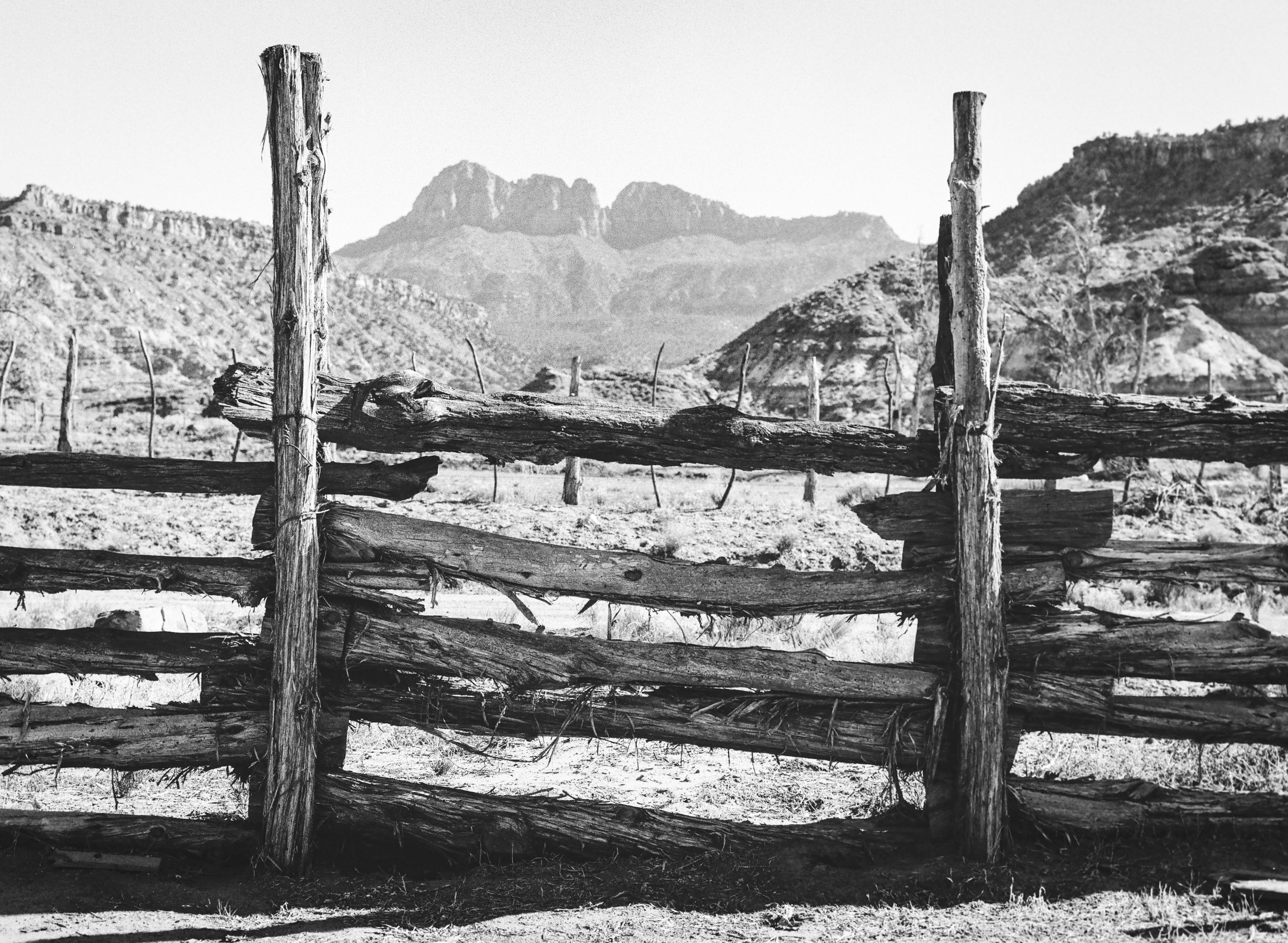 A rustic wooden fence stands in the foreground of a dry, rugged landscape, with mountains visible in the distant background under a clear sky, capturing the timeless charm of film photography. | Photo by Millworks Film Photography & Prints