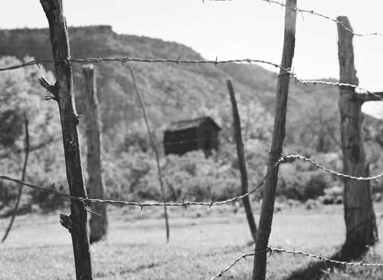 Black and white film photo of a barbed wire fence in the foreground, with an old wooden barn and distant hills softly blurred behind. The scene exudes rural charm, capturing rustic beauty through timeless film photography. | Photo by Millworks Film Photography & Prints