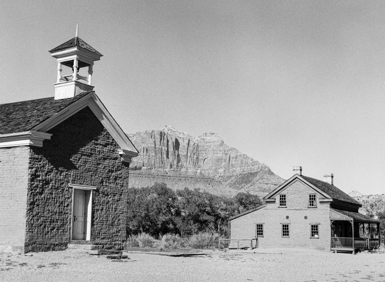 A black and white film photo captures a historic brick schoolhouse and a second brick building, with mountains and trees in the background under a clear sky. | Photo by Millworks Film Photography & Prints