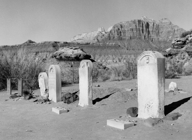 Four old gravestones stand in a barren, desert cemetery with rocky terrain and rugged mountains in the background under a clear sky, captured through evocative film photography. | Photo by Millworks Film Photography & Prints