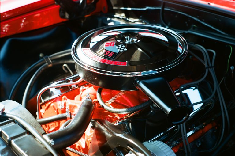 Close-up film photo of a classic Chevrolet 396 cubic inch V8 engine with chrome and black air cleaner cover, red engine block, and visible hoses and wires in a cars engine bay. | Photo by Millworks Film Photography & Prints