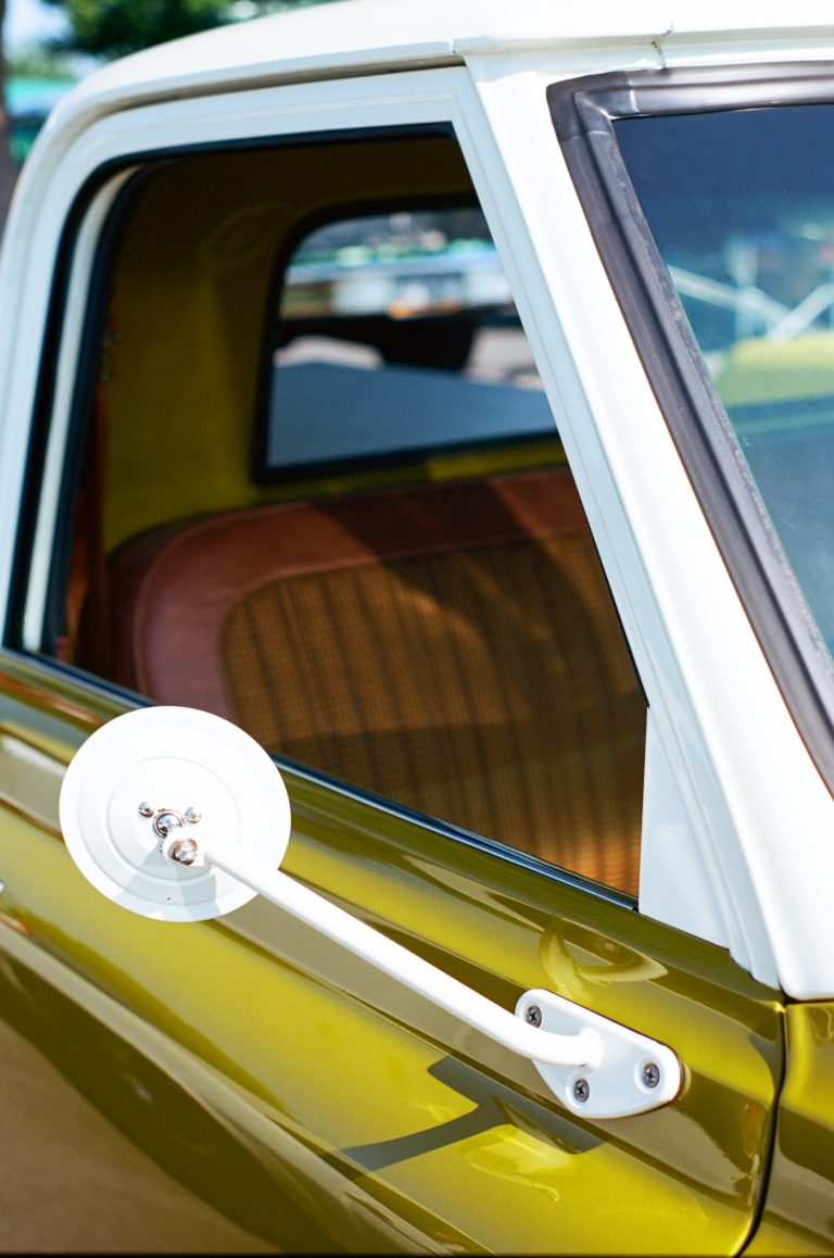 A close-up film photo of an open window on a vintage car with olive green paint, showing a round white side mirror and brown plaid-patterned seats inside. | Photo by Millworks Film Photography & Prints