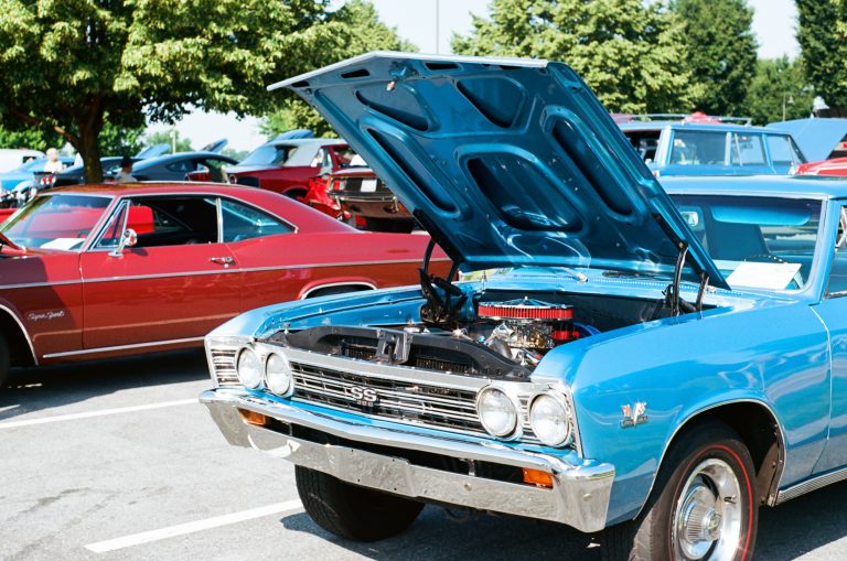 A blue classic Chevrolet SS muscle car with its hood open is parked at a car show next to a red vintage Chevrolet, captured in stunning professional photography. Other classic cars and green trees are visible in the background. | Photo by Millworks Film Photography & Prints