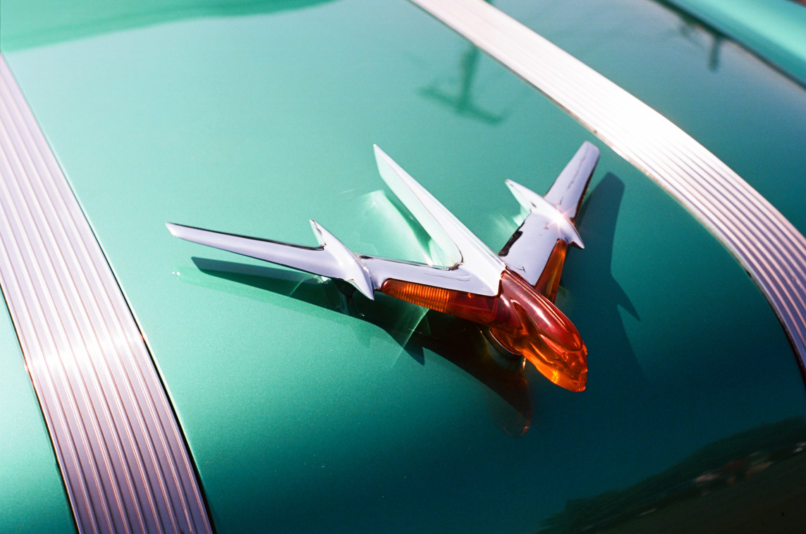 A close-up film photo of a green classic car hood features a shiny chrome hood ornament shaped like an airplane with an orange accent, alongside metal stripes. | Photo by Millworks Film Photography & Prints