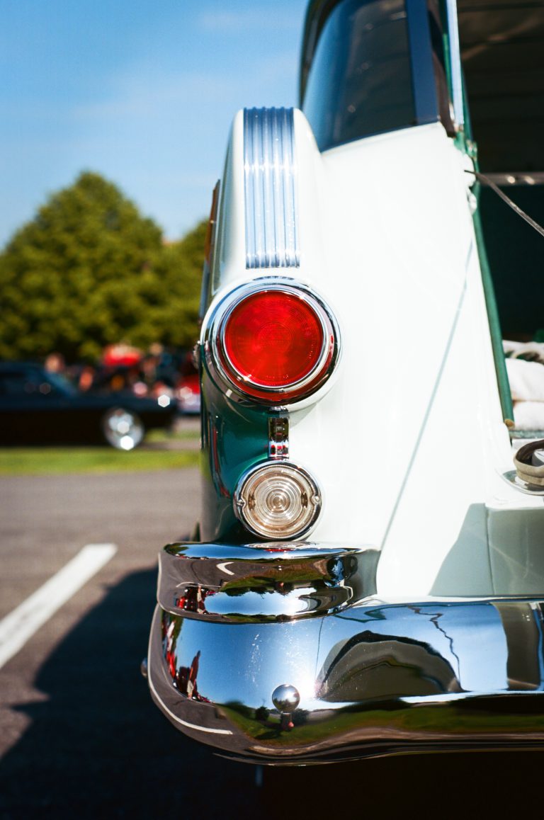 Close-up of the rear tail light and chrome bumper of a vintage white car parked outdoors, captured in rich detail with film photography, with trees and other cars blurred in the background on a sunny day. | Photo by Millworks Film Photography & Prints