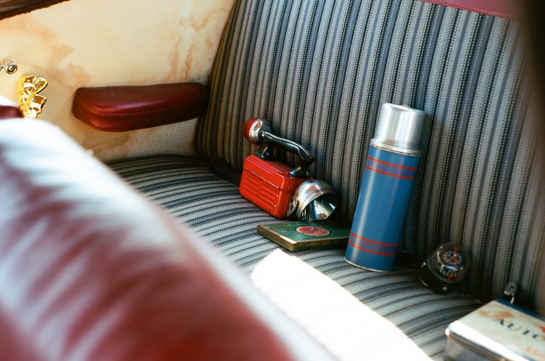 Vintage striped car seat with a blue thermos, red flashlight, metal box, compass, and black device rests on the fabric. The seat and armrest are trimmed in red leather—a scene captured through film photography. | Photo by Millworks Film Photography & Prints