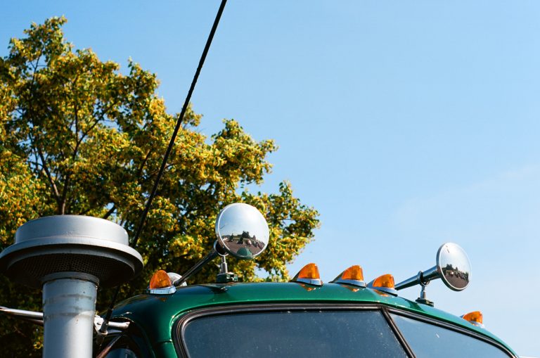 A close-up film photo of the top of a green truck with orange lights, side mirrors, and a metal exhaust pipe, set against a clear blue sky and leafy tree in the background. | Photo by Millworks Film Photography & Prints