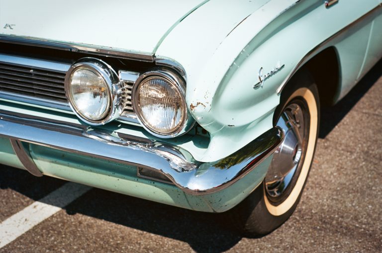 Close-up of the front left side of a vintage light blue car, captured as a film photo, showing round headlights, chrome bumper, and part of a whitewall tire. The car’s paint is slightly chipped near the headlights. | Photo by Millworks Film Photography & Prints