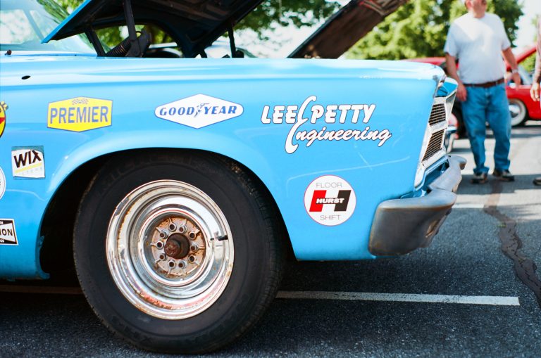 A close-up film photo of the front left side of a vintage blue race car with various decals, including “LEE PETTY engineering.” The hood is open, and a person stands blurred in the background. | Photo by Millworks Film Photography & Prints