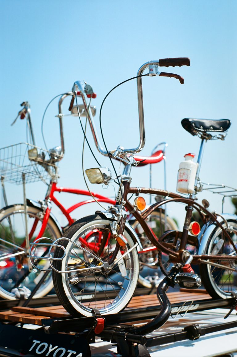 Two vintage bicycles, one red and one brown, are mounted on a car roof rack under a clear blue sky. Captured in the timeless style of film photography, the bikes feature tall handlebars, baskets, and a water bottle on one frame. | Photo by Millworks Film Photography & Prints