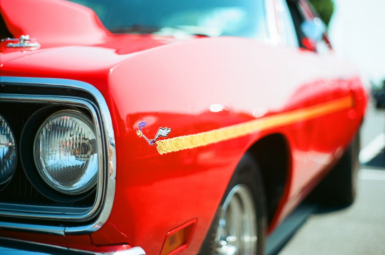 Close-up of a shiny red classic car, captured with film photography, showing the front left headlight, chrome bumper, yellow stripe, and a small cartoon Road Runner decal on the fender, with the background softly out of focus. | Photo by Millworks Film Photography & Prints