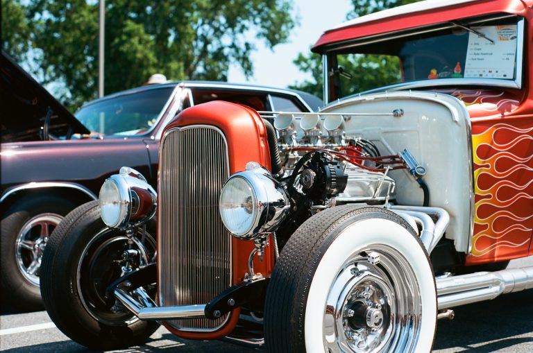 A close-up film photo of a vintage hot rod with a shiny chrome grille, exposed engine, whitewall tires, and flame decals on the side, parked at a car show with another classic car in the background. | Photo by Millworks Film Photography & Prints