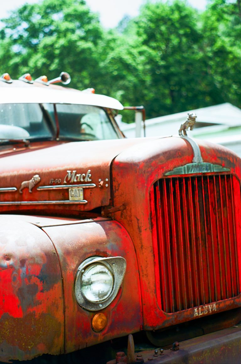 A close-up film photo of an old, rusty red truck with a prominent front grille and headlight. The hood ornament is shaped like a dog, with trees and houses softly blurred in the background, showcasing classic professional photography style. | Photo by Millworks Film Photography & Prints