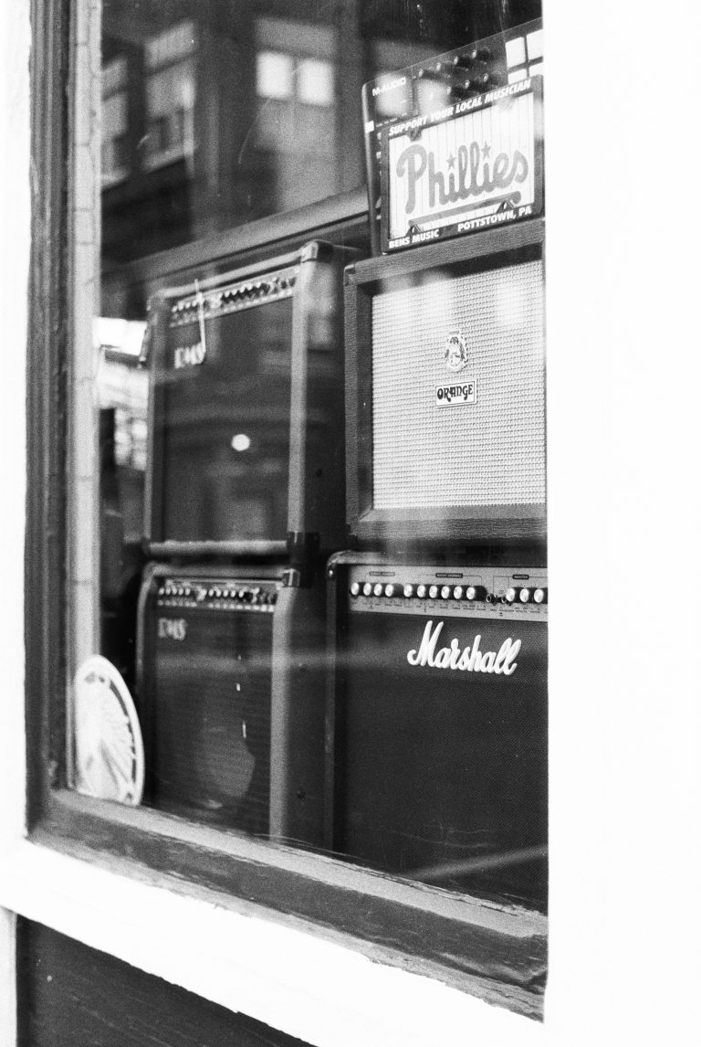 Black-and-white film photography captures a window display of guitar amplifiers, including Marshall and Orange brands, with a Phillies sign and reflections of buildings shimmering in the glass. | Photo by Millworks Film Photography & Prints