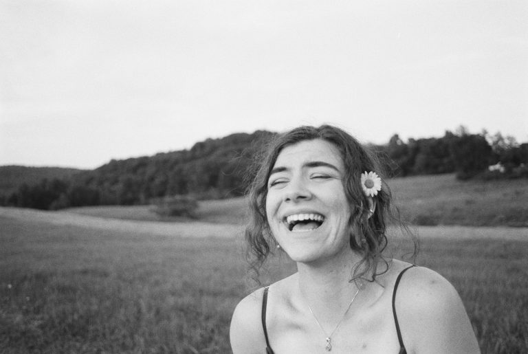 A young woman with wavy hair and a flower tucked behind her ear laughs joyfully in an open field, with trees and hills in the background. The black and white film photo creates a serene, timeless feel. | Photo by Millworks Film Photography & Prints
