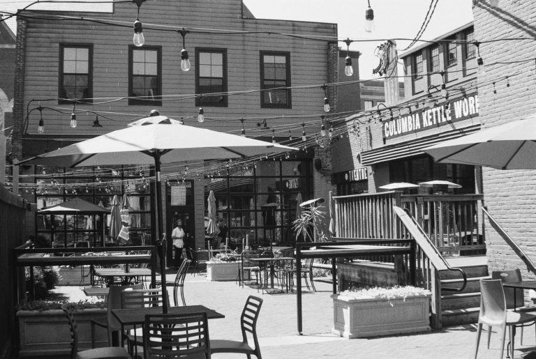 A black and white film photo of an outdoor café with string lights, umbrellas, empty tables and chairs. Two people stand near the entrance. A sign reads Columbia Kettle Works. The urban setting feels relaxed and timeless. | Photo by Millworks Film Photography & Prints