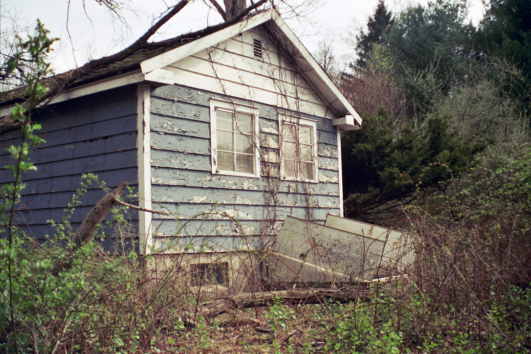 An old, weathered blue house with peeling paint sits among overgrown bushes and weeds. Captured in the nostalgic style of film photography, a fallen tree branch and broken door hint at years of neglect and abandonment. | Photo by Millworks Film Photography & Prints