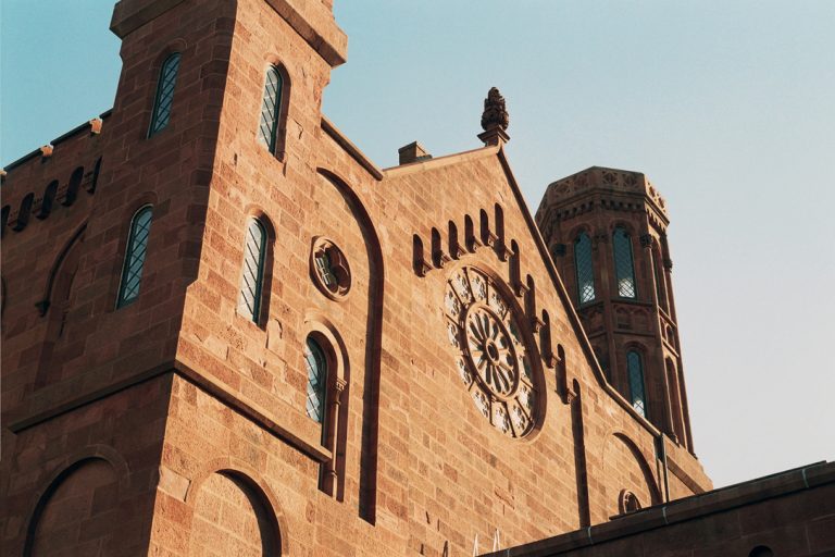 A close-up view of a historic stone building with arched windows, a circular rose window, and a tall octagonal tower under a clear blue sky, captured with the timeless charm of film photography. | Photo by Millworks Film Photography & Prints