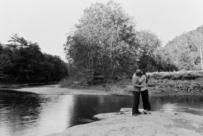 A couple stands on rocks by a river, embracing and kissing, surrounded by trees and natural scenery in this timeless black and white film photo. | Photo by Millworks Film Photography & Prints