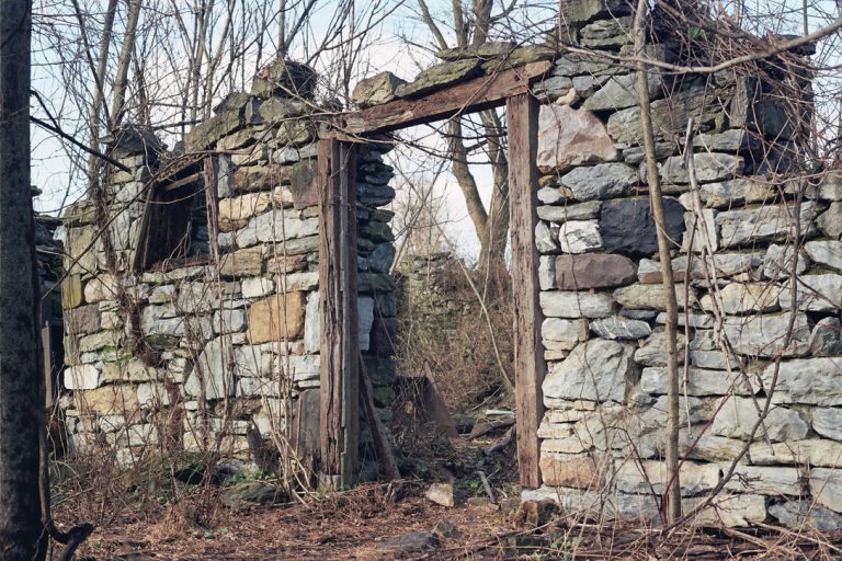 Old, crumbling stone wall with a doorway stands among leafless trees. Captured in atmospheric film photography, the scene’s brown leaves, branches, and overgrowth evoke an abandoned building deep within a forested area. | Photo by Millworks Film Photography & Prints