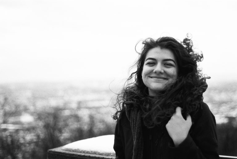 A young woman with curly hair smiles at the camera outdoors on a cold day, wearing a coat and scarf, captured in stunning film photography against a blurred, snowy landscape in the background. | Photo by Millworks Film Photography & Prints