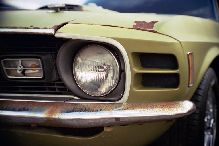 Close-up of the front left headlight and bumper of an old, weathered green car with visible rust and chipped paint, captured in the timeless style of film photography. | Photo by Millworks Film Photography & Prints