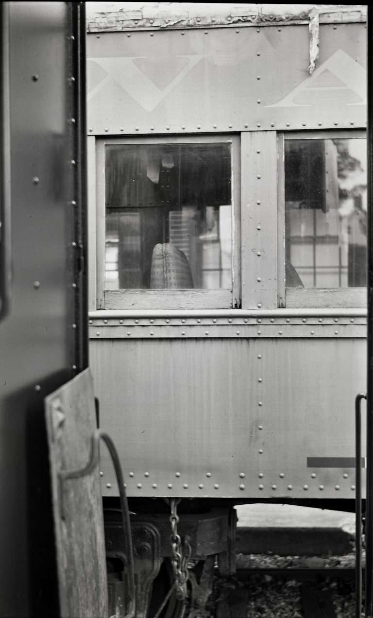 A black-and-white film photo of a train car window shows a person sitting inside with their back to the glass. Framed by another train car in the foreground, this film photography captures a quiet, contemplative moment. | Photo by Millworks Film Photography & Prints