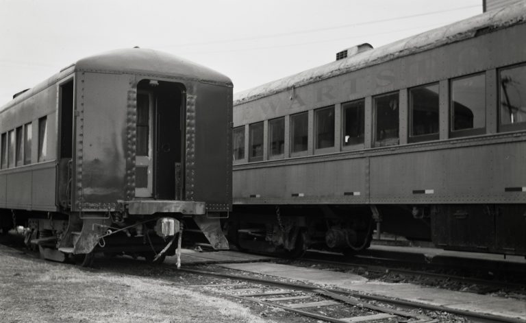 Two vintage train cars are parked side by side on railway tracks; one car’s rear door is open while the other has multiple windows and faded lettering. Captured as a film photo, the quiet scene exudes a nostalgic charm. | Photo by Millworks Film Photography & Prints