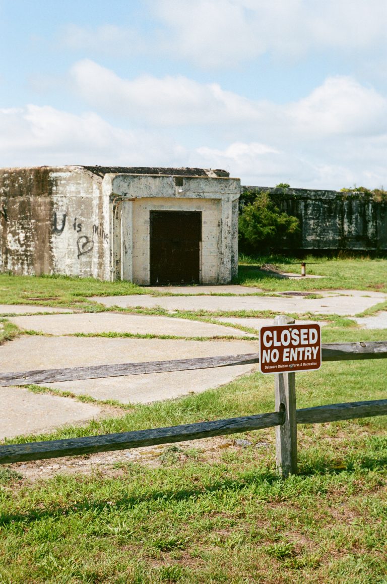 A weathered concrete bunker stands behind a wooden fence with a Closed No Entry sign in front, surrounded by grass and a partly cloudy sky. | Photo by Millworks Film Photography & Prints