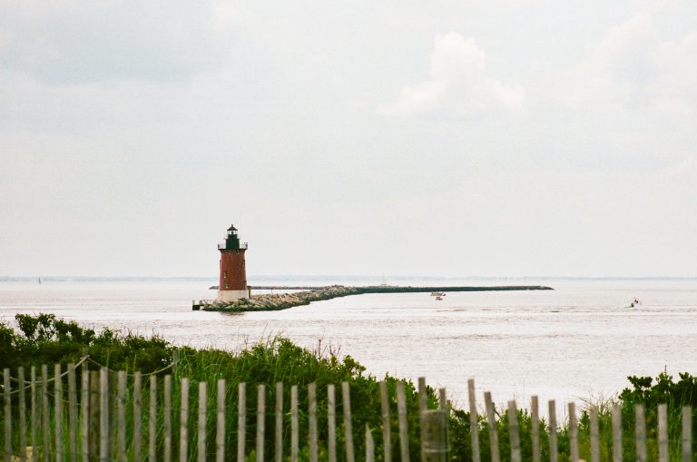 A red brick lighthouse stands at the end of a rocky breakwater, extending into calm water under a cloudy sky. Wooden fencing and green shrubs line the sandy foreground. | Photo by Millworks Film Photography & Prints