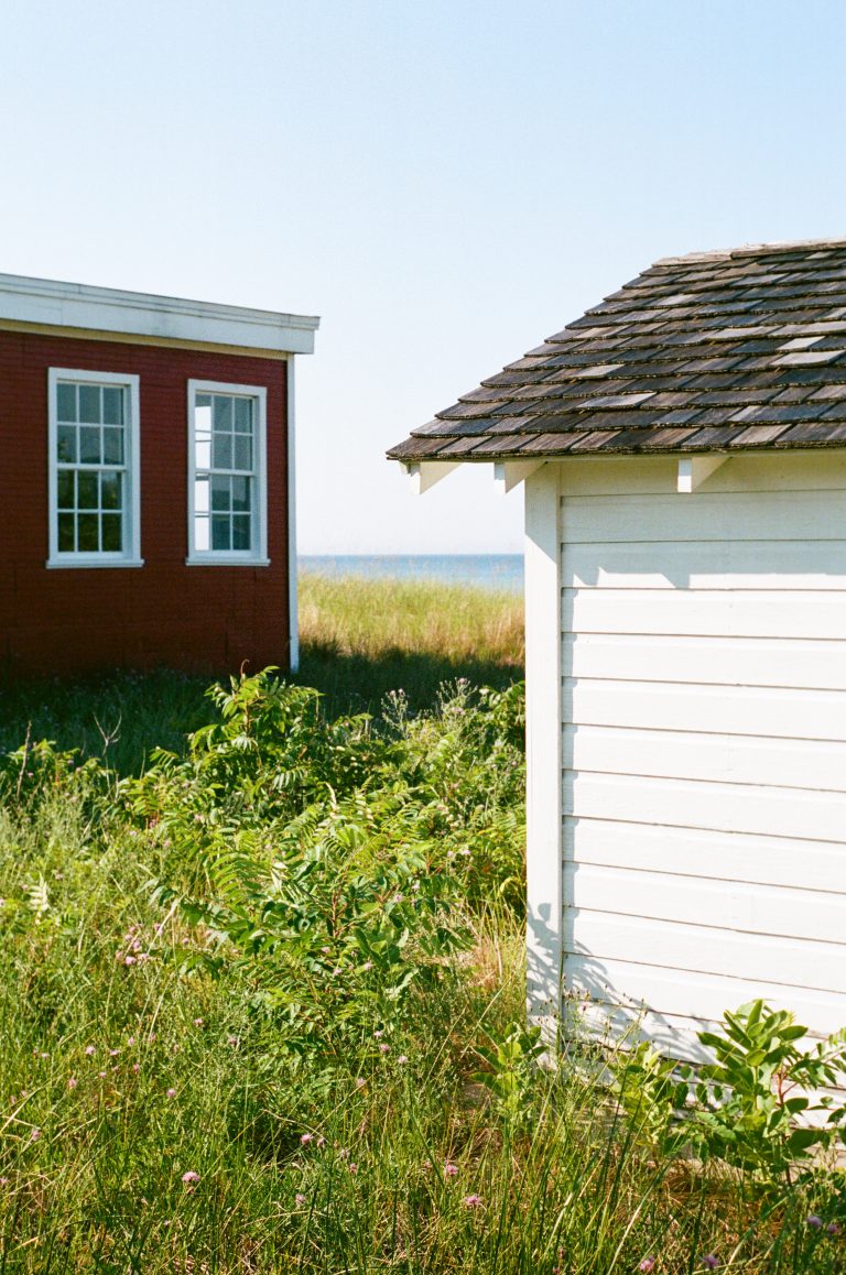 A red building with white-trimmed windows and a white shed sit among tall grass, with the sea and blue sky visible in the background. | Photo by Millworks Film Photography & Prints