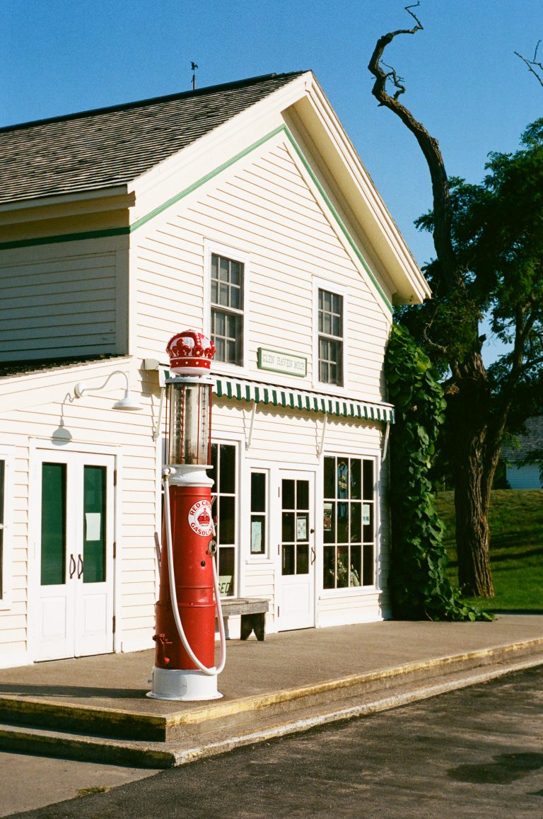 A vintage red gas pump stands on the sidewalk in front of a white, two-story building with green trim and striped awnings, next to a large leafy tree under a clear blue sky. | Photo by Millworks Film Photography & Prints