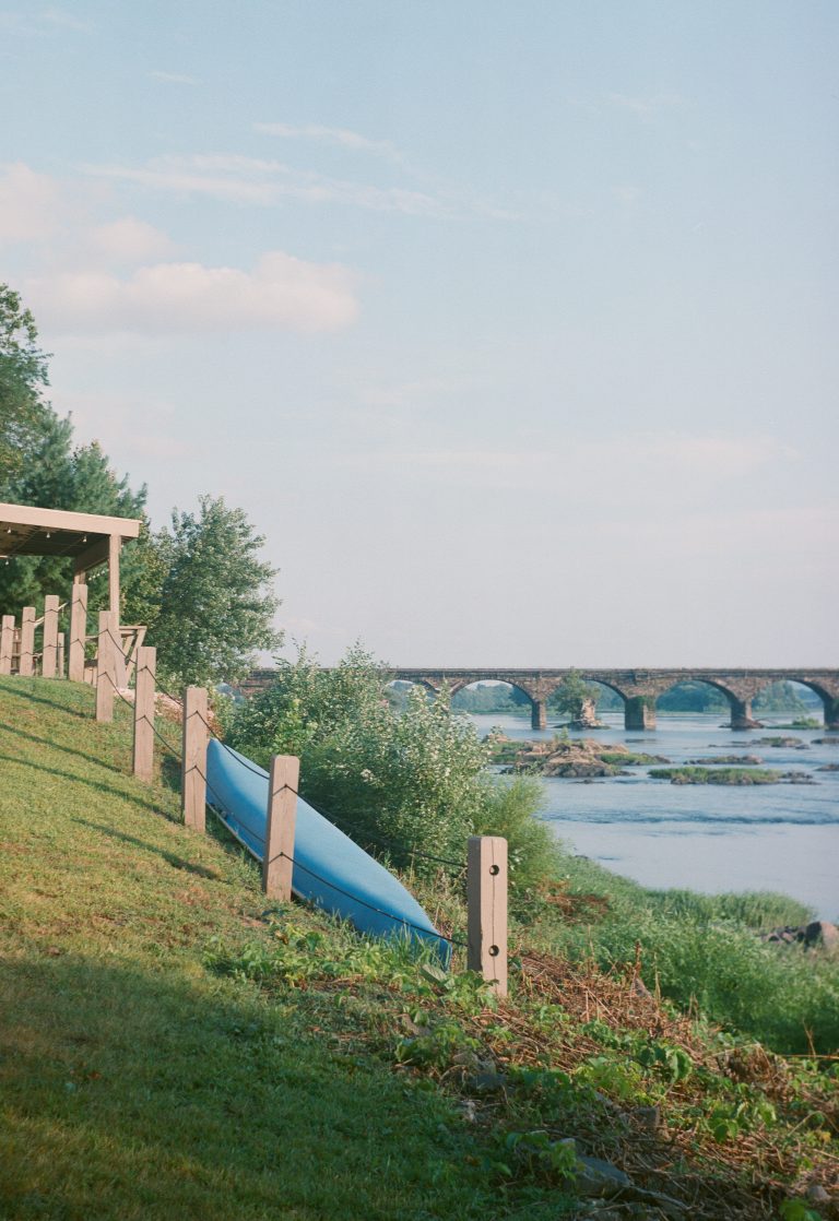 A blue canoe rests upside down on the grassy riverbank next to a wooden fence. A stone arched bridge spans the river in the background under a partly cloudy sky. Trees and tall grass border the peaceful scene. | Photo by Millworks Film Photography & Prints