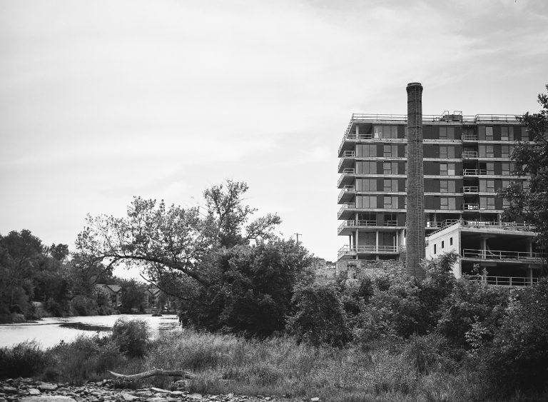 A partially constructed multi-story building with scaffolding stands beside a river, surrounded by trees and dense vegetation, under a cloudy sky in black and white. | Photo by Millworks Film Photography & Prints