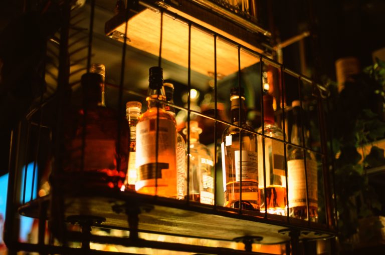 A selection of liquor bottles sits on a dimly lit bar shelf, enclosed by a metal rack, with warm lighting creating a cozy atmosphere reminiscent of nature-inspired landscapes. | Photo by Millworks Film Photography & Prints