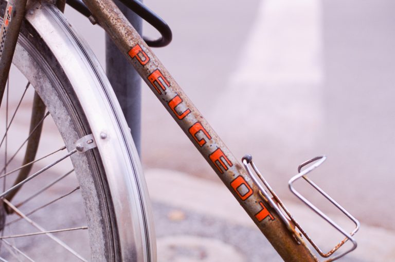 Close-up of a weathered bicycle frame with the word PEUGEOT in orange letters. The image also shows part of the rear wheel, fender, and bottle holder, set against blurred pavement and a crosswalk—urban landscapes meet vintage charm. | Photo by Millworks Film Photography & Prints