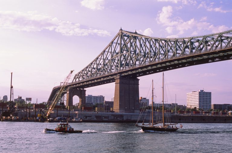 Large steel bridge crossing a river framed by urban landscapes, with a tugboat and sailboat passing underneath; city buildings, cranes, and hints of nature beneath a partly cloudy sky in the background. | Photo by Millworks Film Photography & Prints