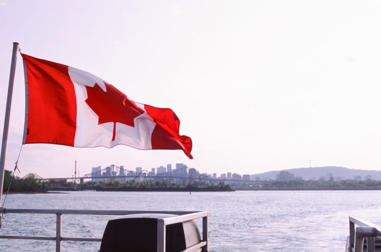 A Canadian flag waves on a boat in the foreground, set against tranquil water and a distant city skyline under a clear sky—an inspiring scene that beautifully blends nature and urban landscapes. | Photo by Millworks Film Photography & Prints