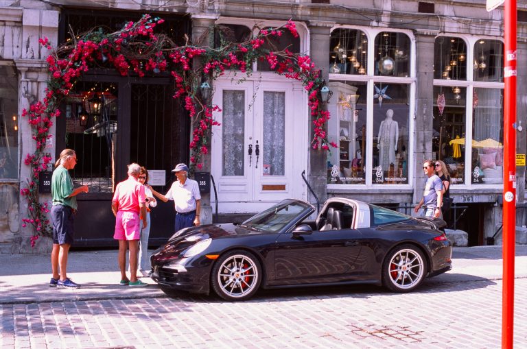 A black convertible sports car is parked on a cobblestone street in front of a building with large windows and pink flowers, blending seamlessly into the vibrant landscapes of urban nature as six people stand nearby, talking and admiring the scene. | Photo by Millworks Film Photography & Prints