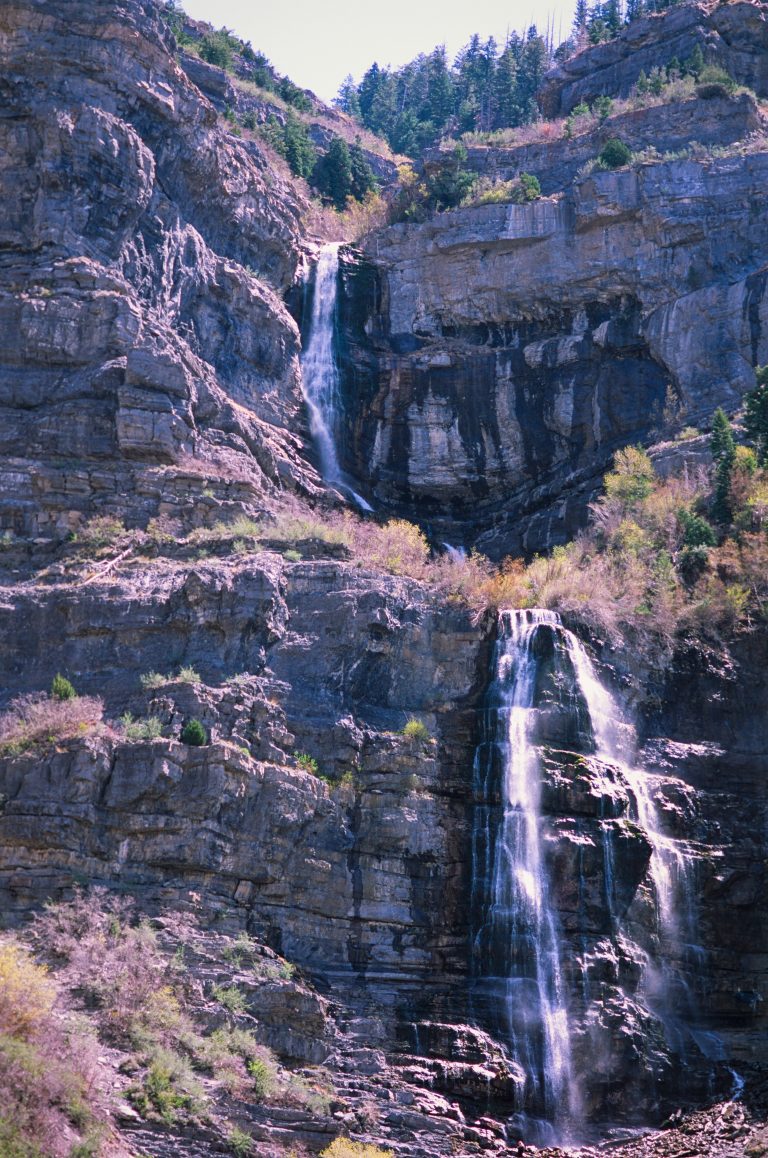Two-tiered waterfall cascades down rugged, rocky cliffs surrounded by sparse trees and vegetation, with sunlight illuminating parts of the rock face and water—an awe-inspiring display of nature’s landscapes. | Photo by Millworks Film Photography & Prints