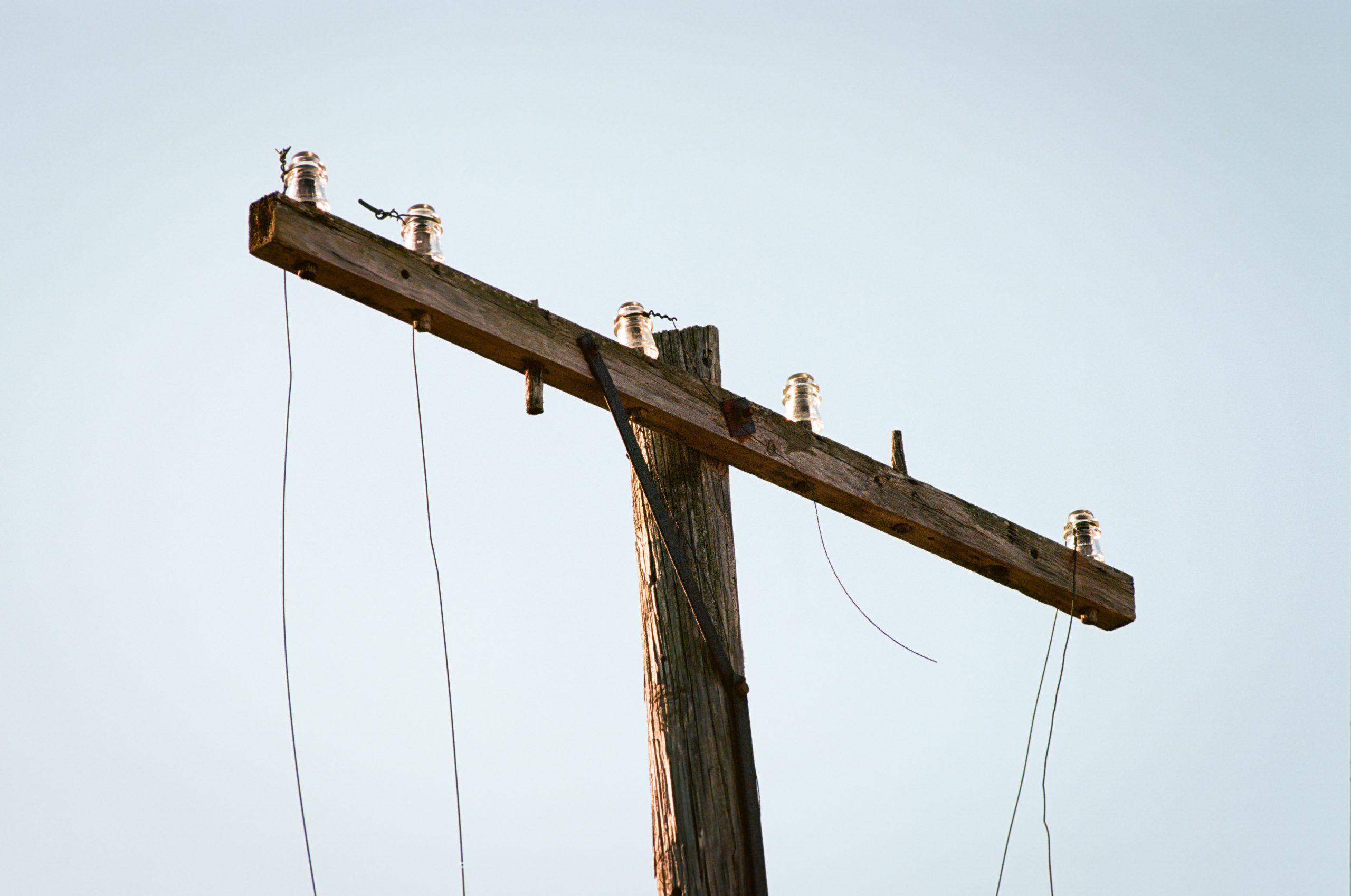 A close-up of an old, weathered wooden utility pole with glass insulators and several loose or broken wires, set against a clear, pale blue sky. | Photo by Millworks Film Photography & Prints