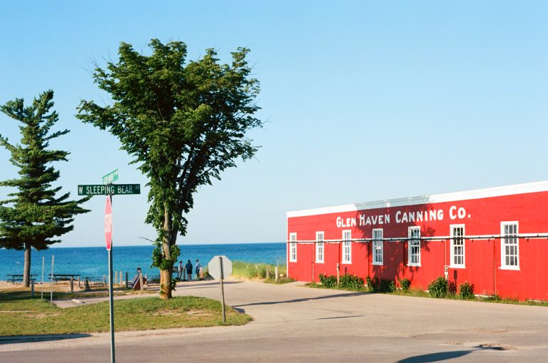 A red building labeled Glen Haven Canning Co. sits by a blue lake under a clear sky, with a street sign for S Sleeping Bear. Trees and a few people are visible near the water in the background. | Photo by Millworks Film Photography & Prints