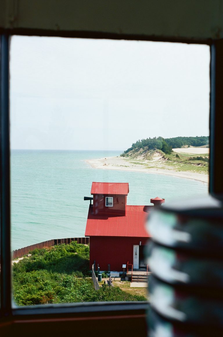 A view through a window shows a red-roofed building by the shore, green bushes, and calm blue-green water stretching toward a sandy peninsula with trees in the distance. | Photo by Millworks Film Photography & Prints