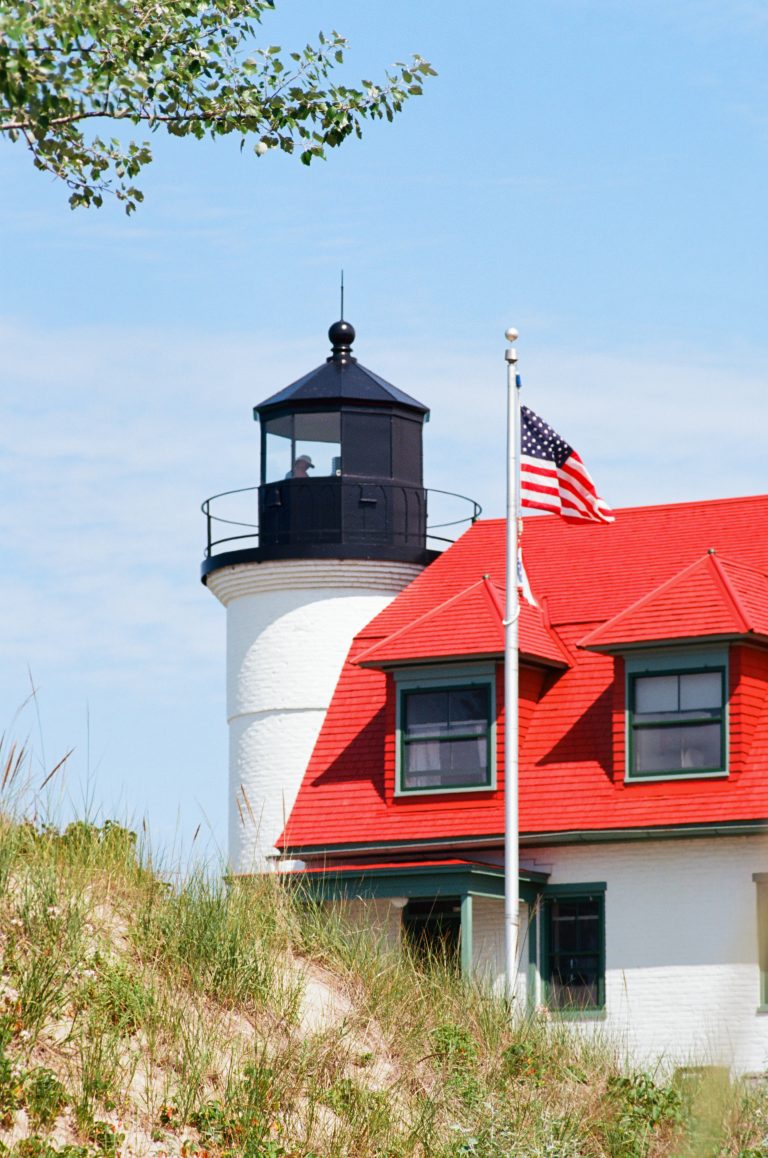 A white lighthouse with a black lantern stands behind a building with a red roof and dormer windows; an American flag waves on a pole in front, with dune grass and a blue sky in the background. | Photo by Millworks Film Photography & Prints
