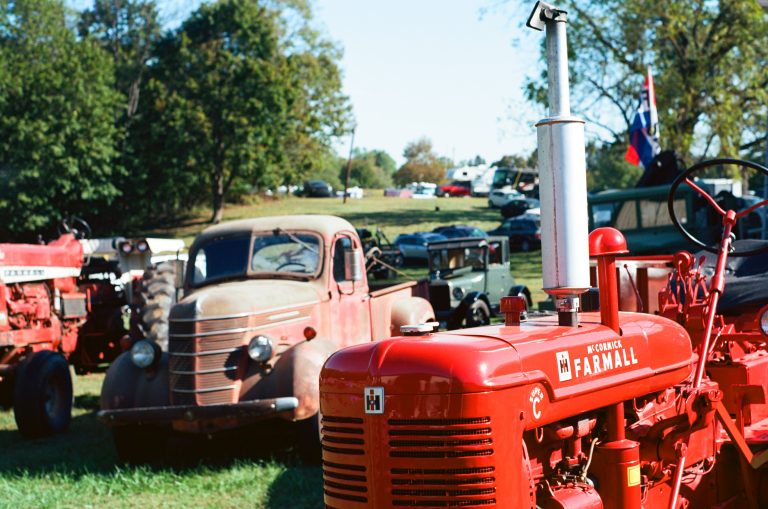 A bright red vintage Farmall tractor is parked on grass beside a rusty old pickup truck at an outdoor event, with more classic vehicles and trees visible in the background under a clear sky. | Photo by Millworks Film Photography & Prints