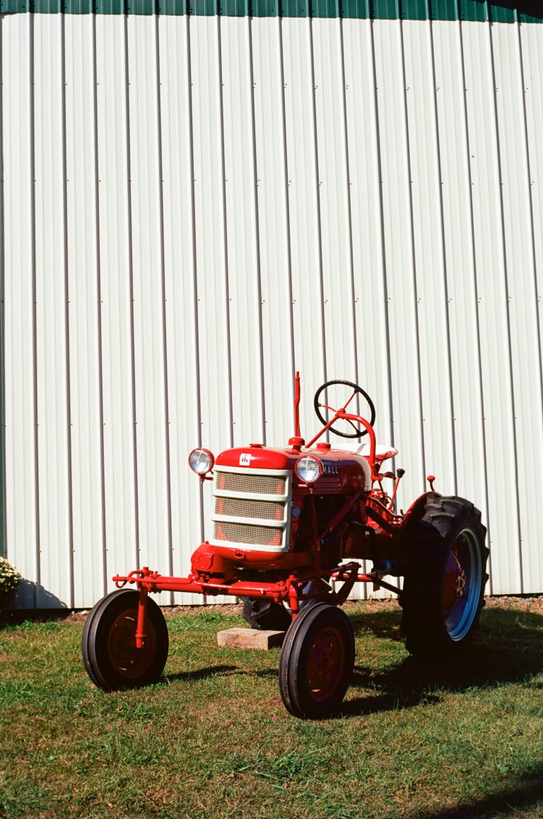 A red vintage tractor is parked on grass in front of a large white metal-sided building on a sunny day. | Photo by Millworks Film Photography & Prints